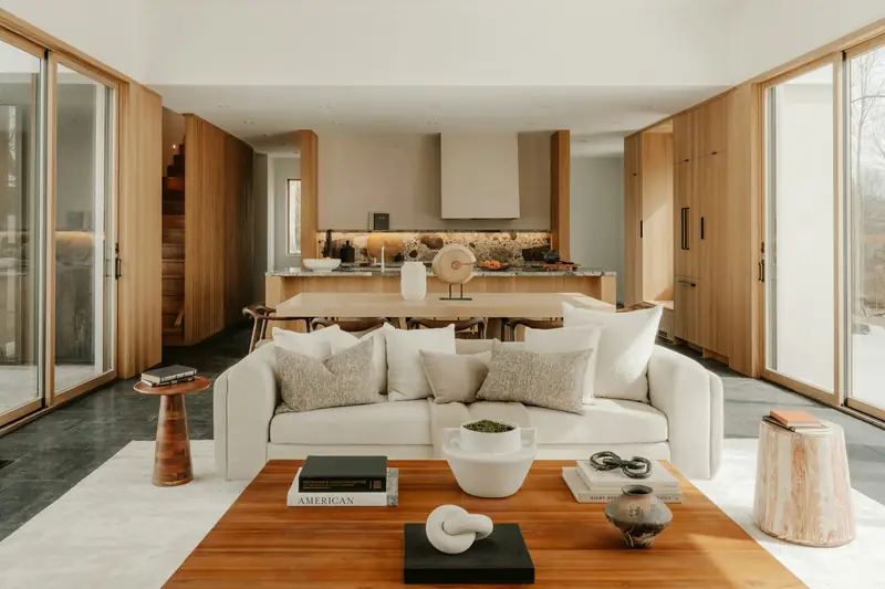 Restrained pale living room with a white sofa, timber kitchen cabinets and stone backsplash, lit through full-height windows.