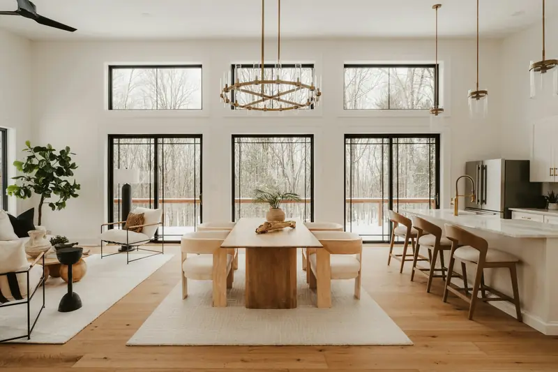 Natural timber dining table, cream chairs and kitchen island in a tall white room overlooking trees through large black-framed windows.
