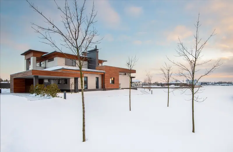 Full view of a modern low-rise timber and light stone house among bare garden trees in snow at soft winter light.