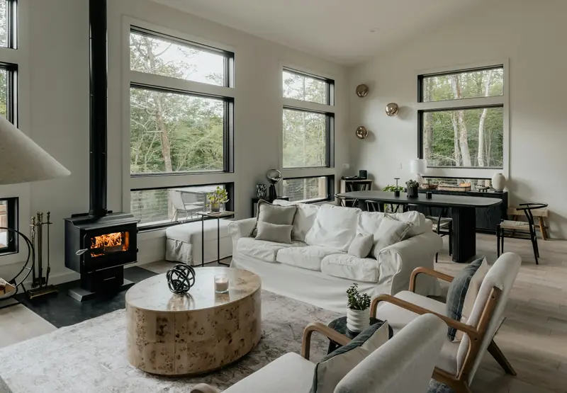 Calm white living room with timber-framed lounge chairs, a stone coffee table and black wood stove beside forest-facing windows.