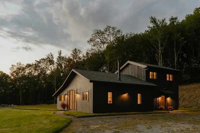 Complete view of a dark timber house with warm lit windows, lawn and a forested hillside under a muted dusk sky.