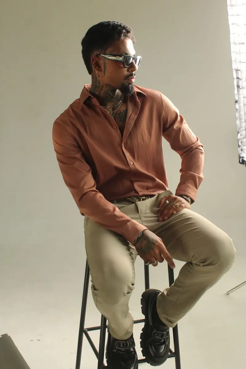 Portrait of a man in a brown shirt and light trousers, seated in a studio.