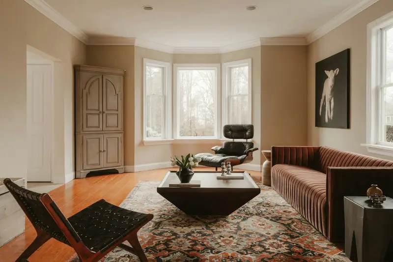 A naturally lit living room photographed by Clay Banks, with bay windows, warm timber flooring, a leather lounge chair and a sculptural coffee table.